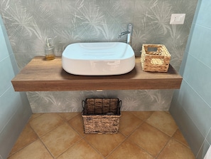 A bathroom with a wooden vanity, a white sink, a faucet, a small glass bottle, and a woven basket. - Residenza Don Alfonso (Somma Vesuviana)
