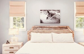 Quarto com cabeceira de vime, uma cama de solteiro com duas almofadas, mesa de cabeceira com luminária e quadro pendurado na parede.
