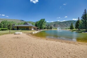 A building with a fenced area, a pond with a fountain, and a mountainous backdrop. - Charming Condo, Near BC/ Vail Resort, BBQ (Avon)