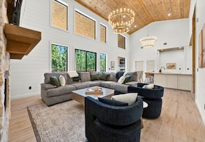 A spacious living room with a sectional sofa, a coffee table, and a fireplace. There are large windows allowing natural light and views of the outdoors. - Children’s Playset | Hot Tub | Games | Sleeps 16 (Broken Bow)
