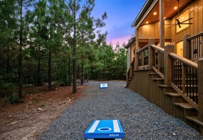 A GoSports shuffleboard setup in a wooded area with a wooden building and stairs in the background. - Children’s Playset | Hot Tub | Games | Sleeps 16 (Broken Bow)