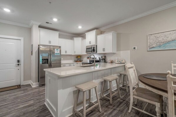 A modern kitchen with white cabinetry, a stainless steel refrigerator, a microwave, and a sink. There is a wooden dining table with chairs. - Stunning&Serene Golf Resort Condo (Calabash)