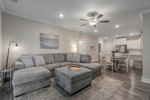 A modern living room with a sectional sofa, a dining area with a table and chairs, a kitchen with white cabinets, and a ceiling fan. - Stunning&Serene Golf Resort Condo (Calabash)