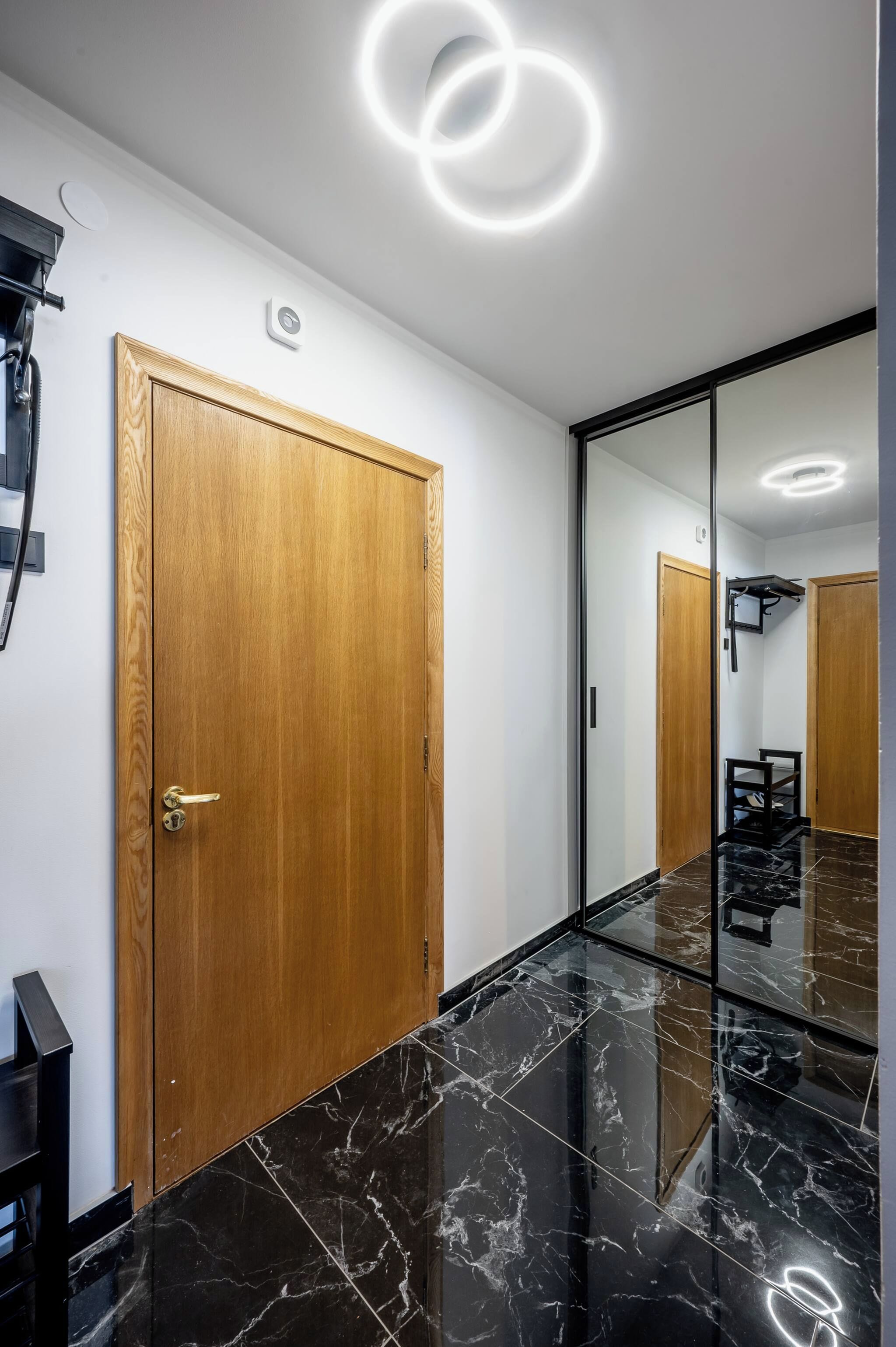 A hallway with a wooden door, a black marble floor, and a large mirror reflecting the door and a ceiling light fixture.