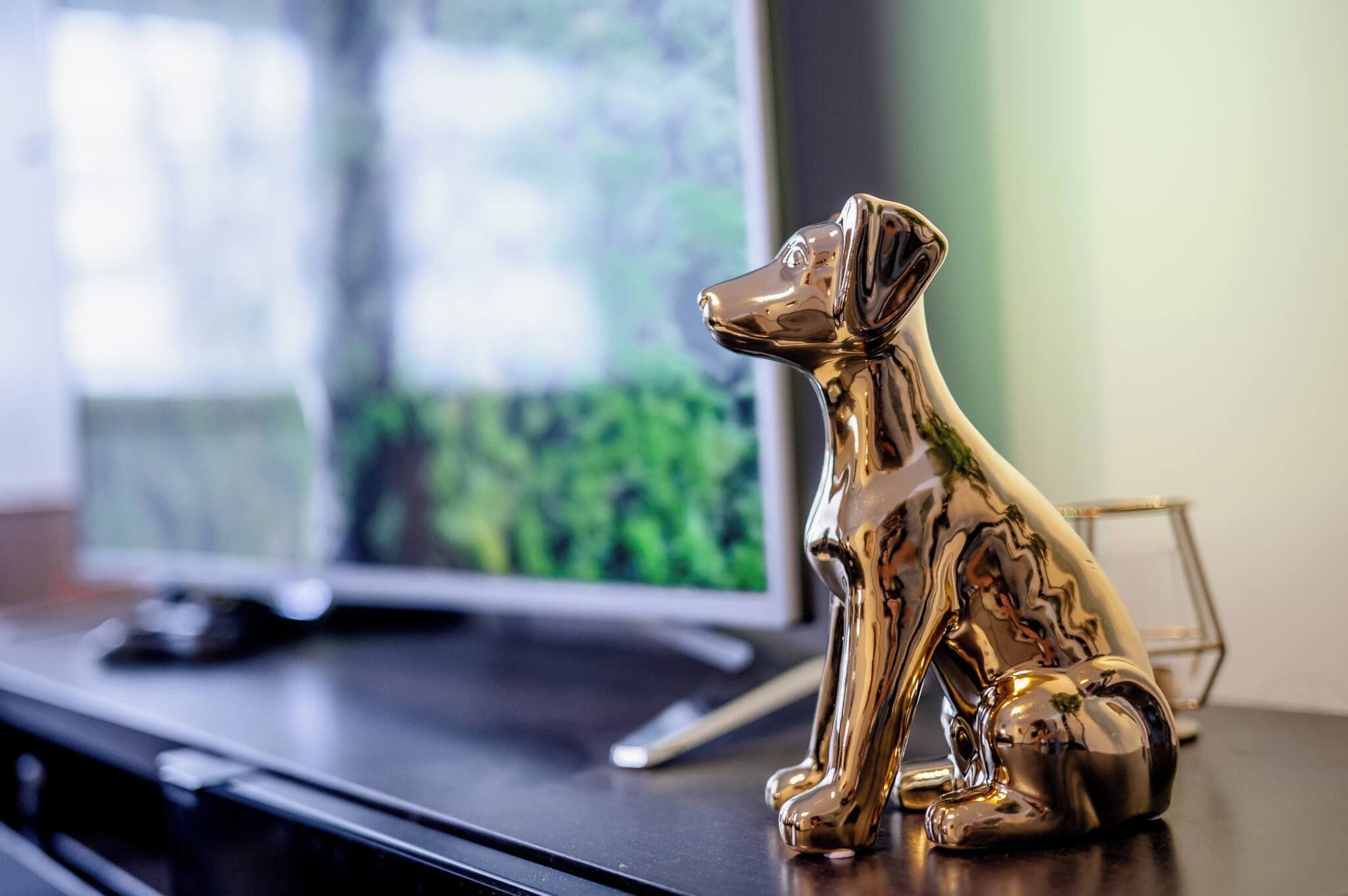 Eine glänzende, metallische Hundfigur auf einer ebenen Fläche, vermutlich einem Tisch, vor verschwommenem Hintergrund mit Fenster und Fernseher.