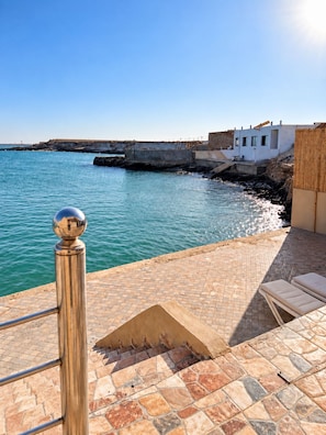 A coastal terrace with a stone floor, a metal railing, and a view of the sea and a white building.