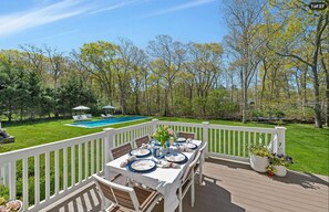 Elevated deck with outdoor dining overlooking a lawn, pool, and surrounding trees. - Hamptons Retreat | Private Pool & 3BR Escape (East Quogue)