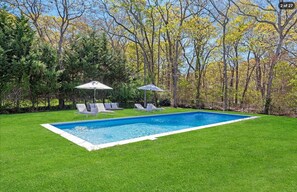 A rectangular in-ground swimming pool on a broad lawn surrounded by woods. - Hamptons Retreat | Private Pool & 3BR Escape (East Quogue)