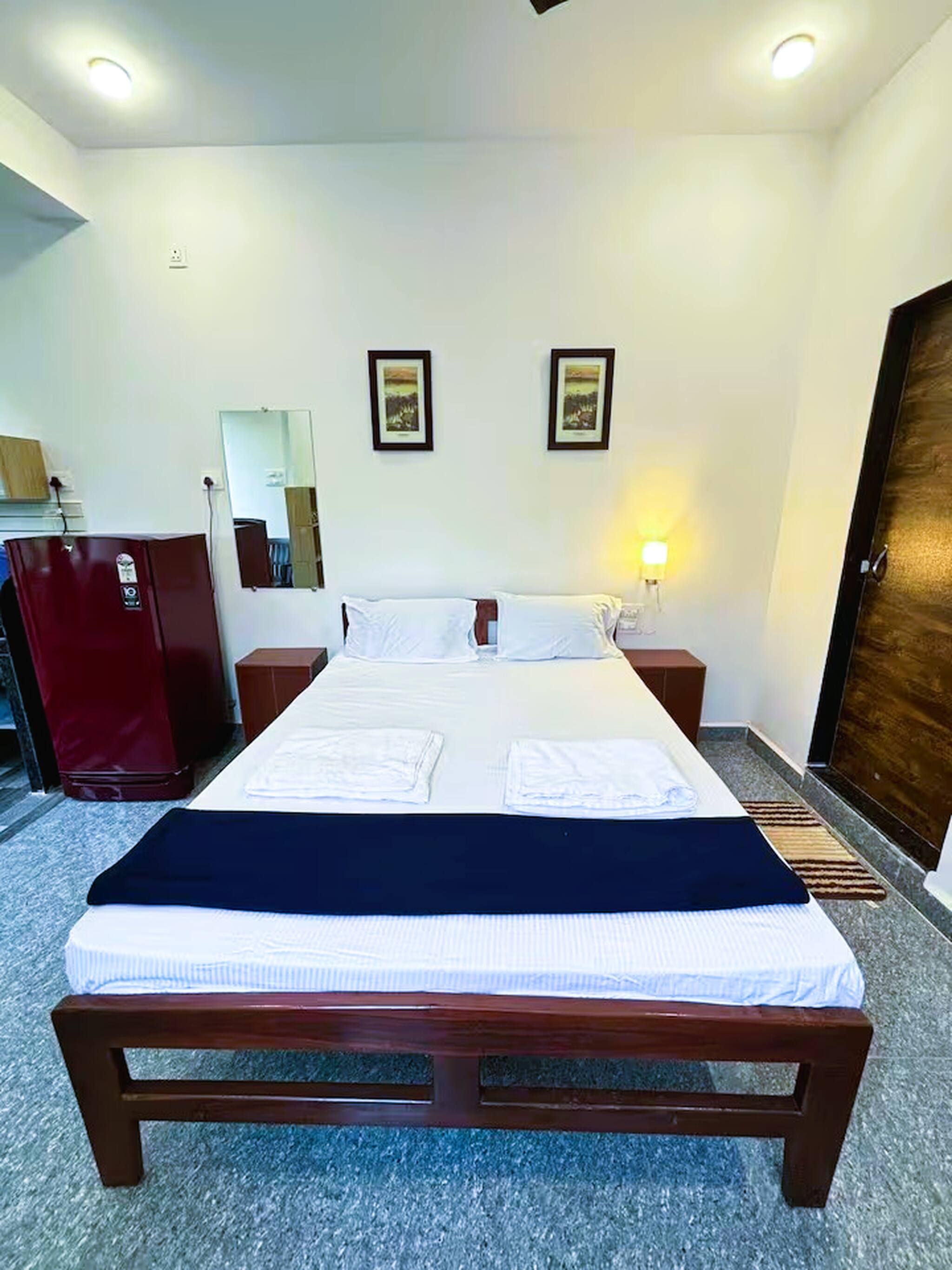 A neatly made bed with white linens and a dark headboard, a wooden nightstand with a lamp, a red suitcase, and a wooden door.