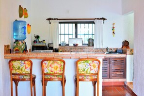 A kitchen with a water dispenser, a coffee maker, and a window with curtains. There are three chairs with floral upholstery. - Above the Gardens. Beyond Expectations - Villa Rosamaria, Pet-Friendly (Manzanillo)