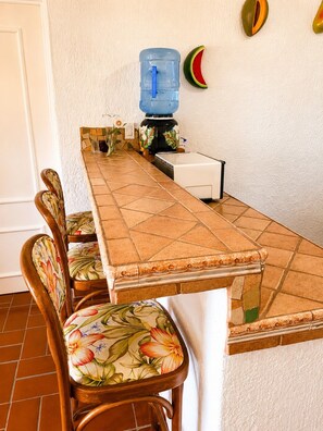 A dining area with a long table, chairs with floral upholstery, a water dispenser, and a microwave. - Above the Gardens. Beyond Expectations - Villa Rosamaria, Pet-Friendly (Manzanillo)