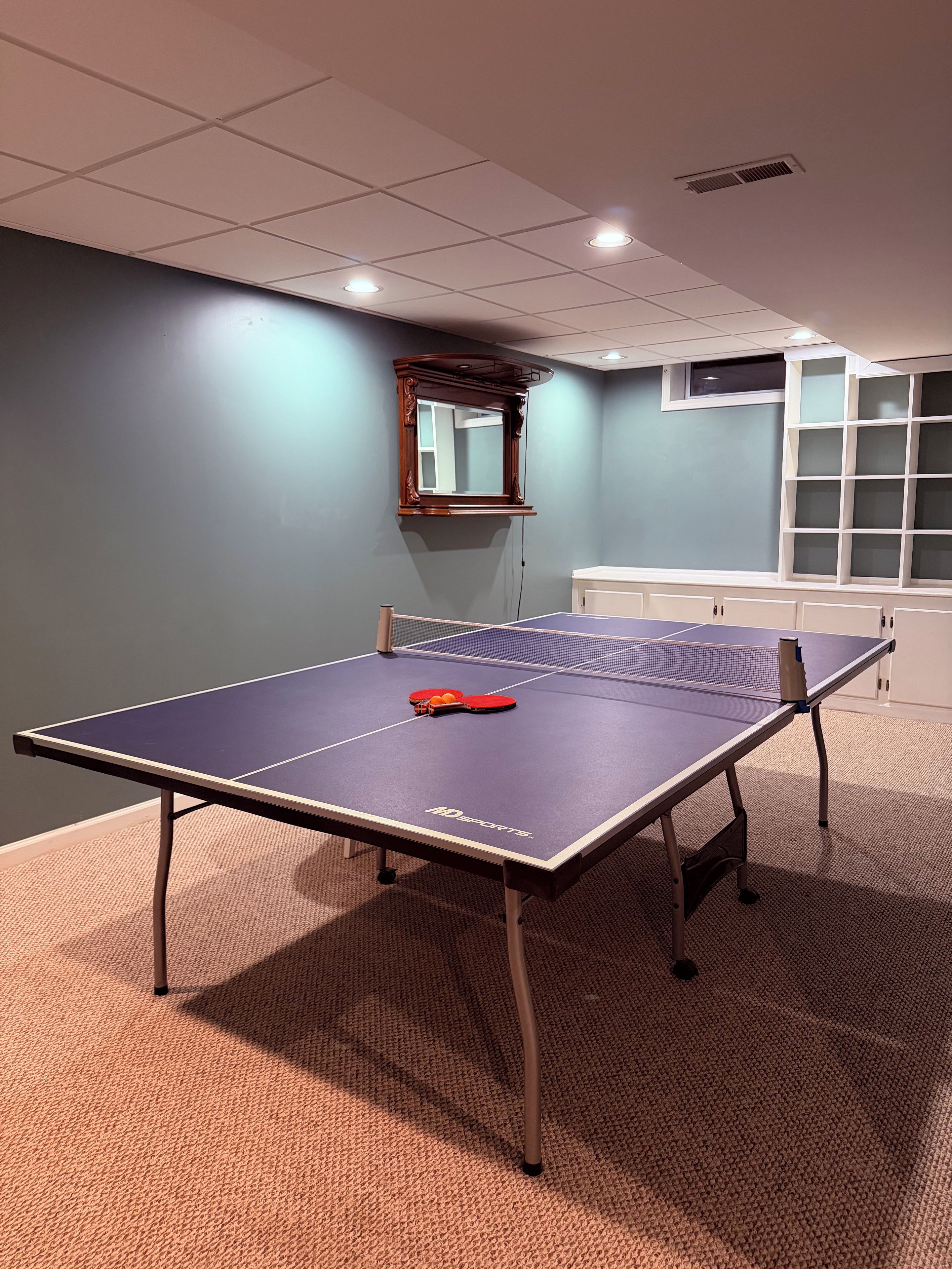 Ein Tischtennistisch mit Schlägern und Ball, ein Spiegel und ein Schrank in einem Raum mit Teppichboden.