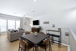 A modern living room with a sofa, a dining table with chairs, a TV mounted on the wall, and a staircase with a railing. - San Antonio Ranch Loft | 2BR/2BA Garage + Pool (San Antonio)