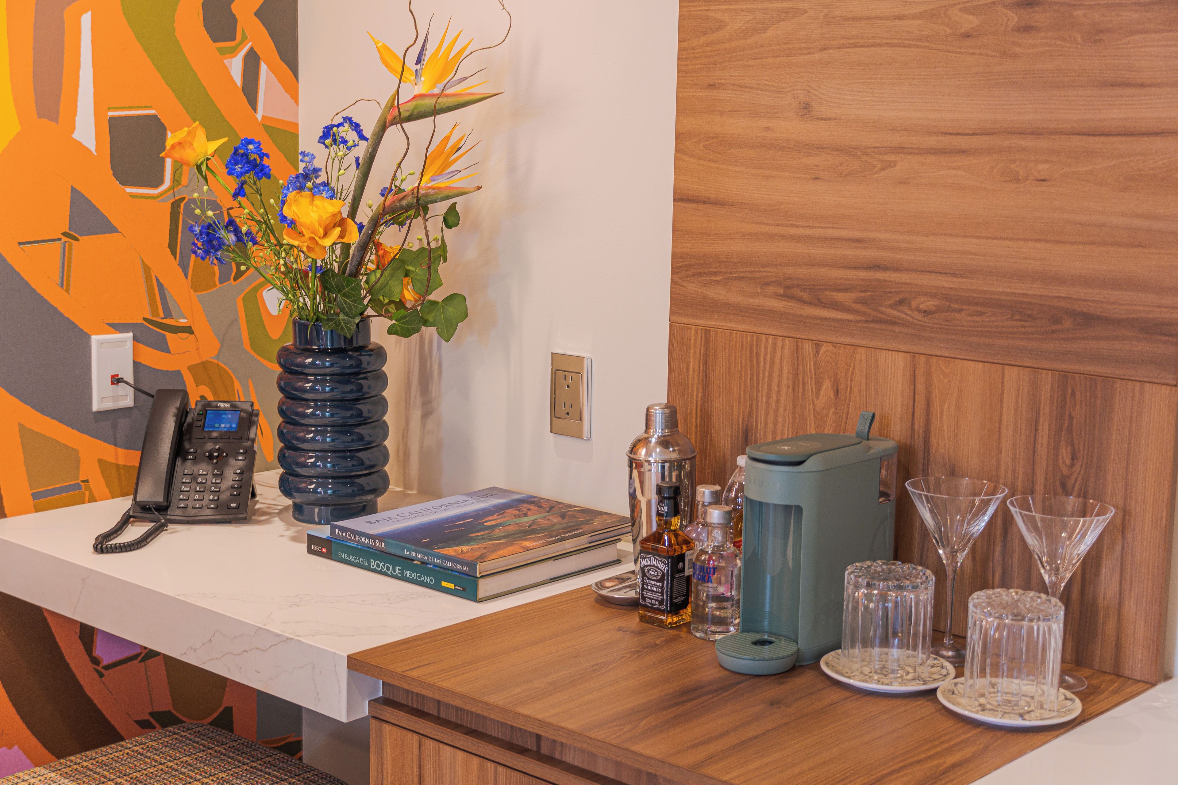 A wooden desk with a phone, books, a vase with flowers, a cocktail shaker, and cocktail glasses.