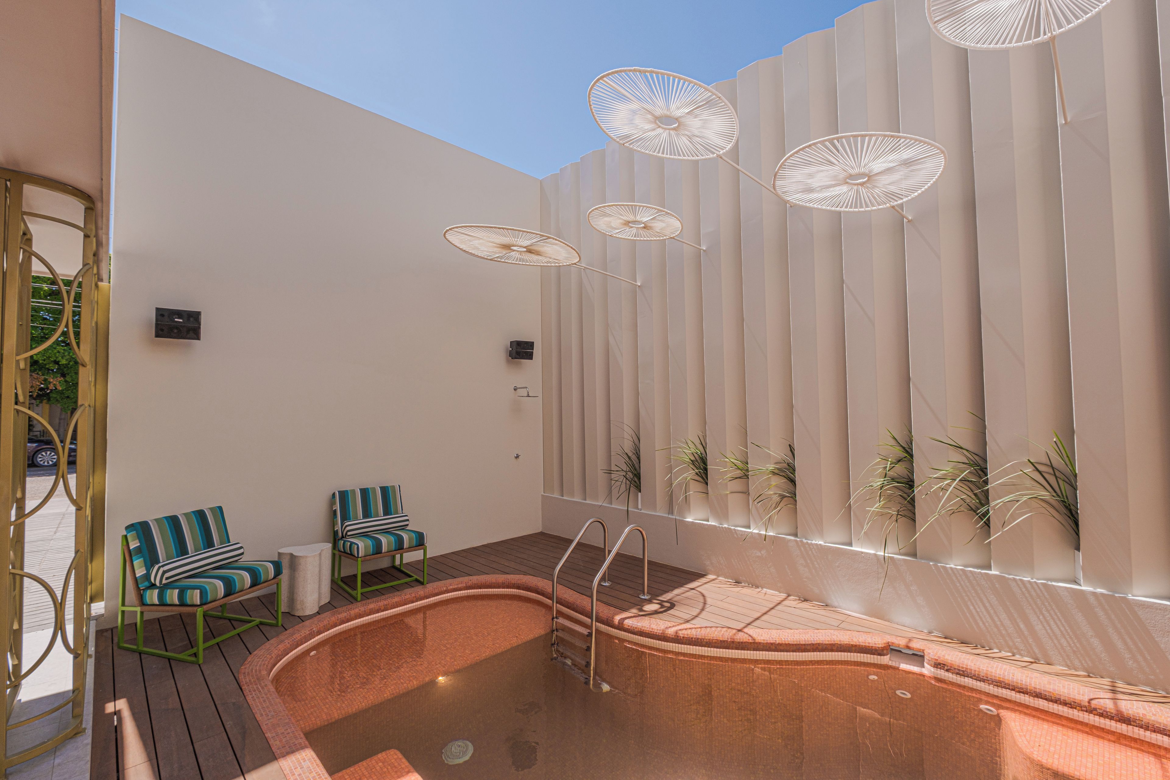 A spa area with a hot tub, two striped chairs, and a wall with vertical slats and hanging umbrellas.