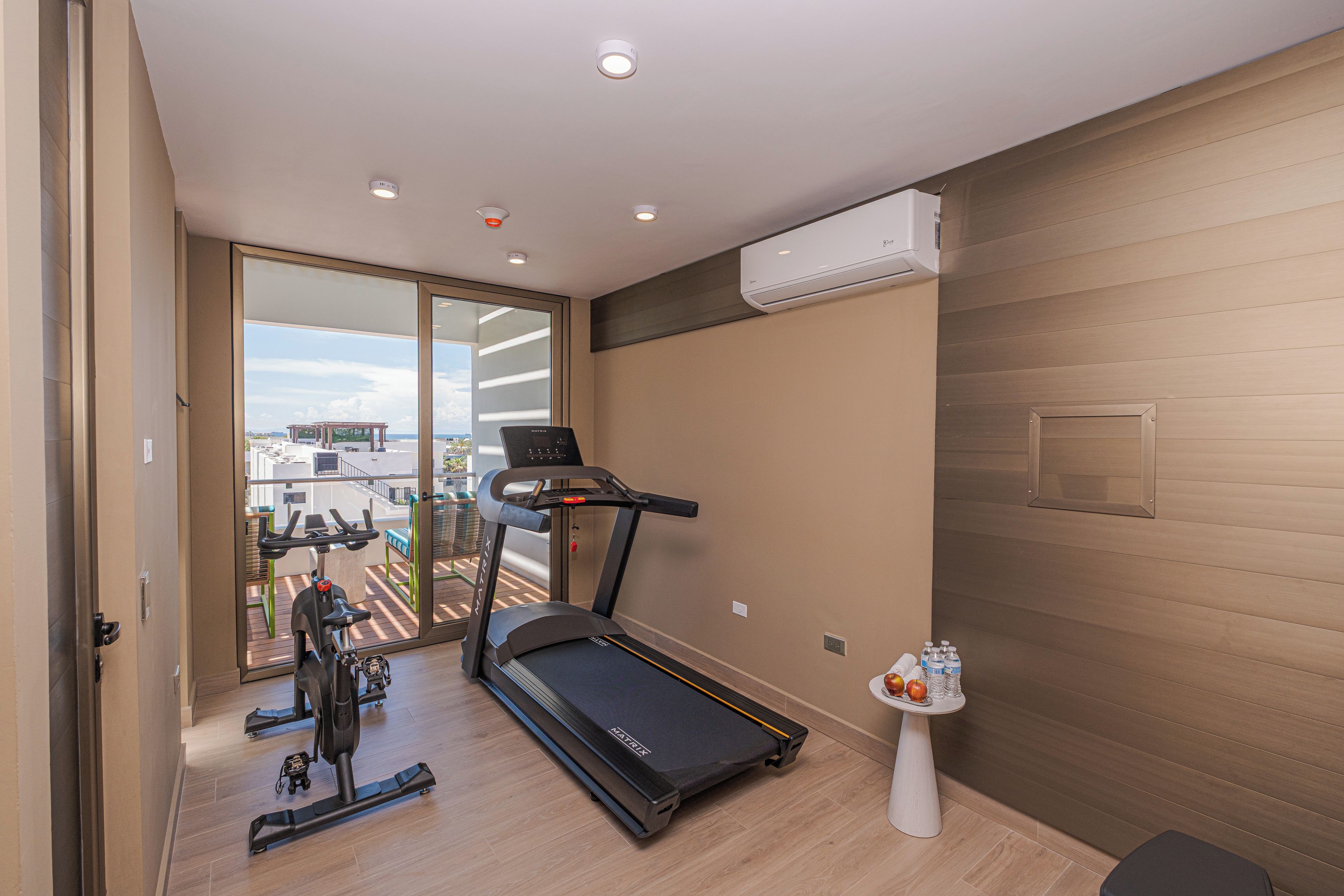 Un gimnasio moderno con cinta de correr, bicicletas estáticas y una pequeña mesa con frutas y botellas de agua.