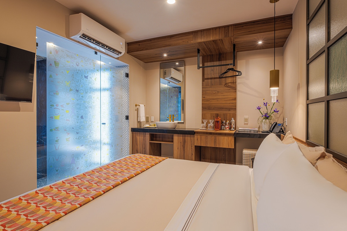 A hotel room with a bed, a bathroom, a vanity with a mirror, and a hanging light fixture.