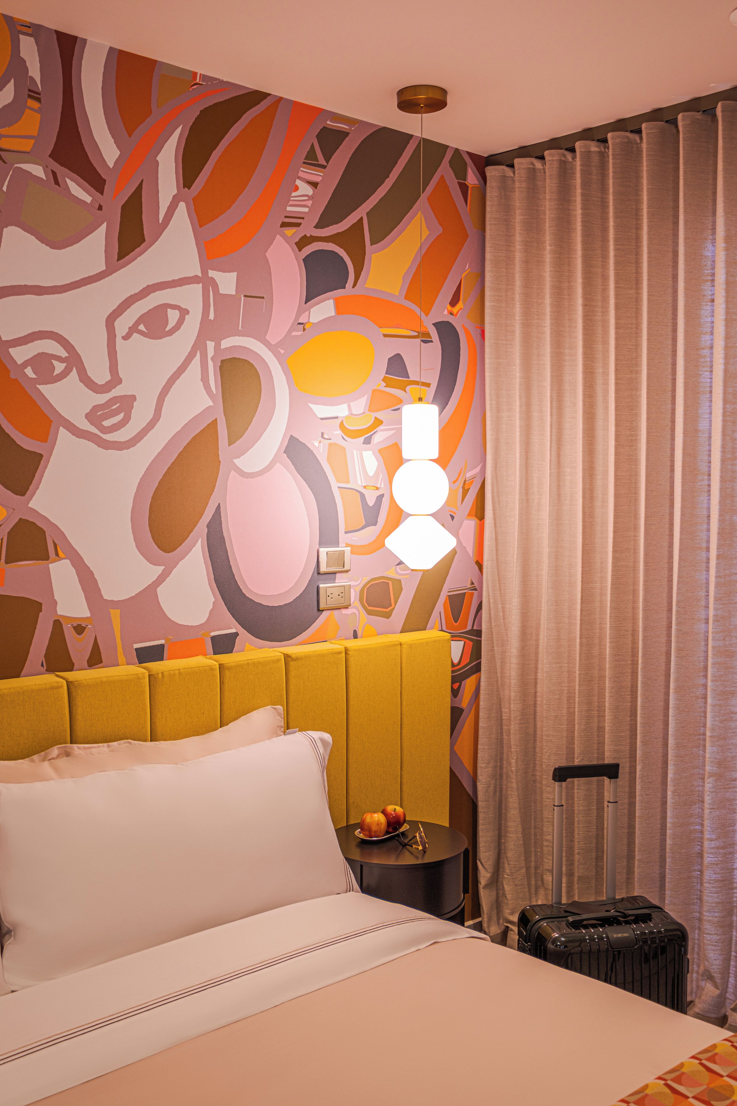 A hotel room with a bed, a headboard, a nightstand, a lamp, a suitcase, and a curtain.