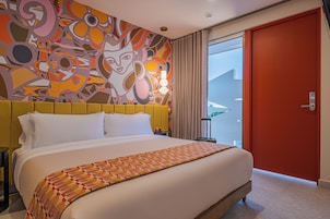 A hotel room with a large bed, a colorful abstract wall mural, a red door, and a view of the outside.