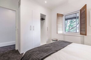 A bedroom with a bed, a wardrobe, a window with shutters, and a small stool. - By the Bay Elegance A Laid back Urban Retreat (Brighton East)