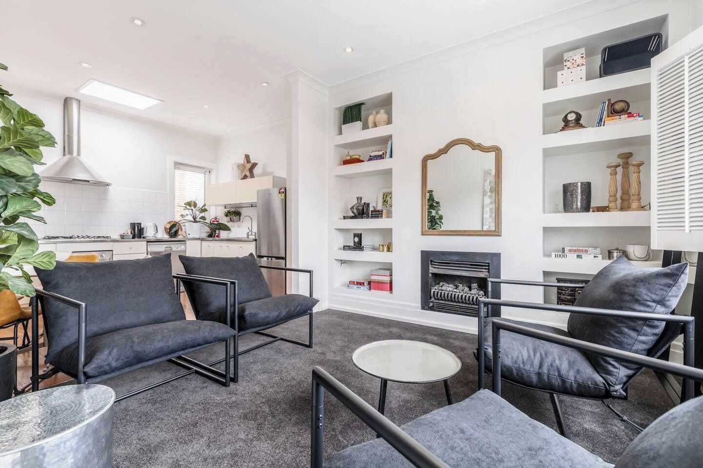 A modern living room with a sofa, armchairs, and a coffee table. There is a fireplace with a mirror above it and shelves with decorative items.