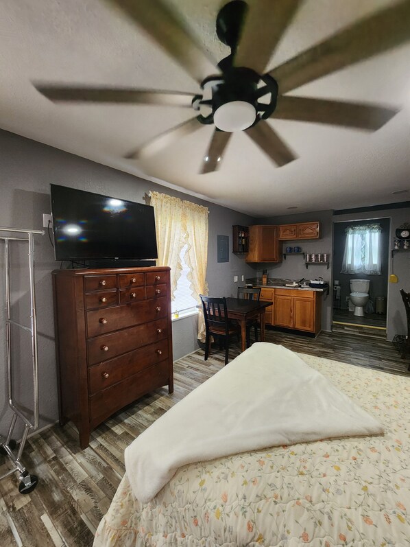 A hotel room with a bed, a dresser, a television, a ceiling fan, a kitchen area, and a bathroom.