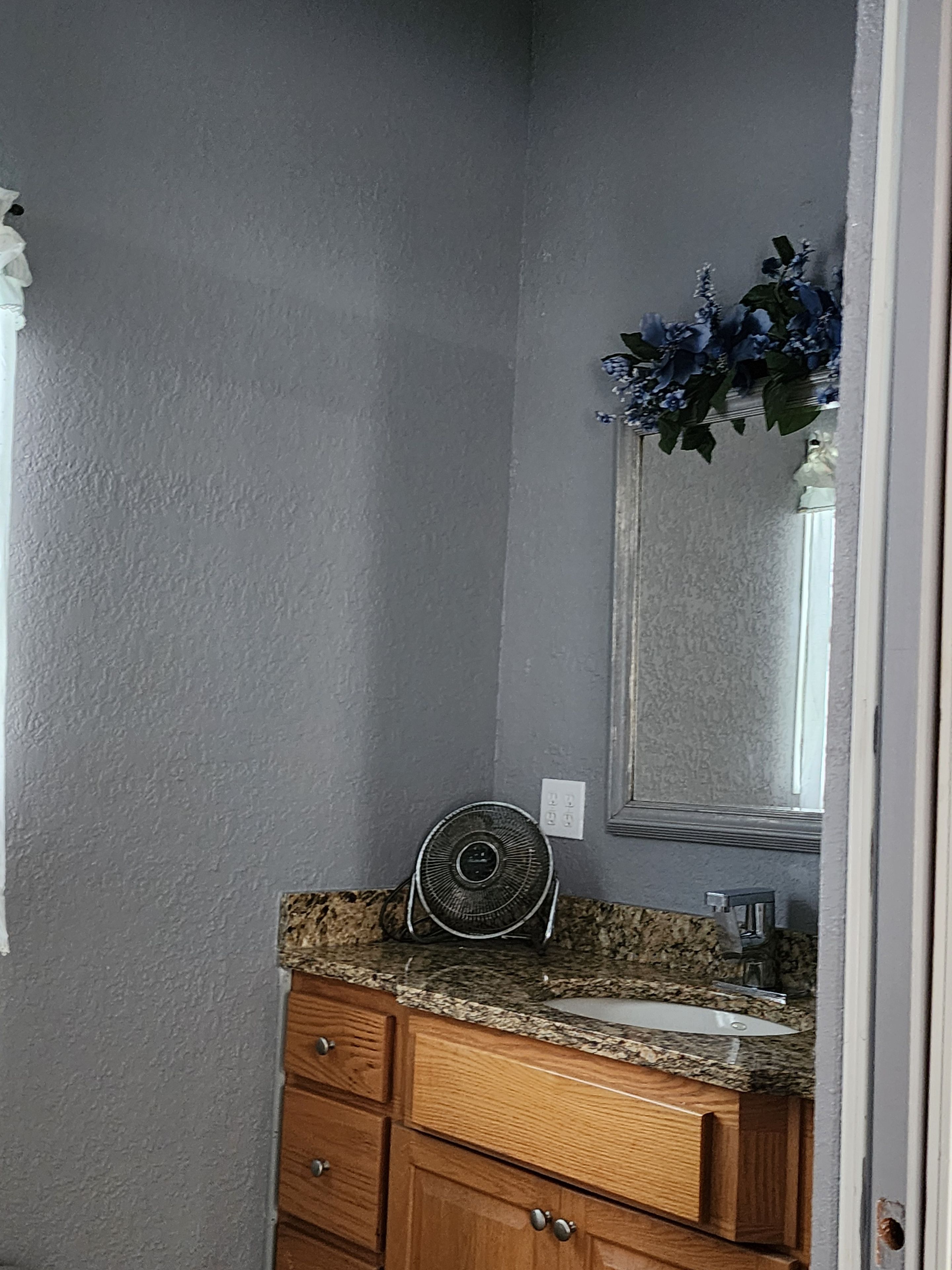 Een badkamer met een granieten aanrechtblad, houten kastjes, een spiegel en een ventilator bevestigd aan de muur.