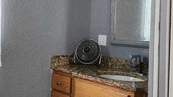 Een badkamer met een granieten aanrechtblad, houten kastjes, een spiegel en een ventilator bevestigd aan de muur.