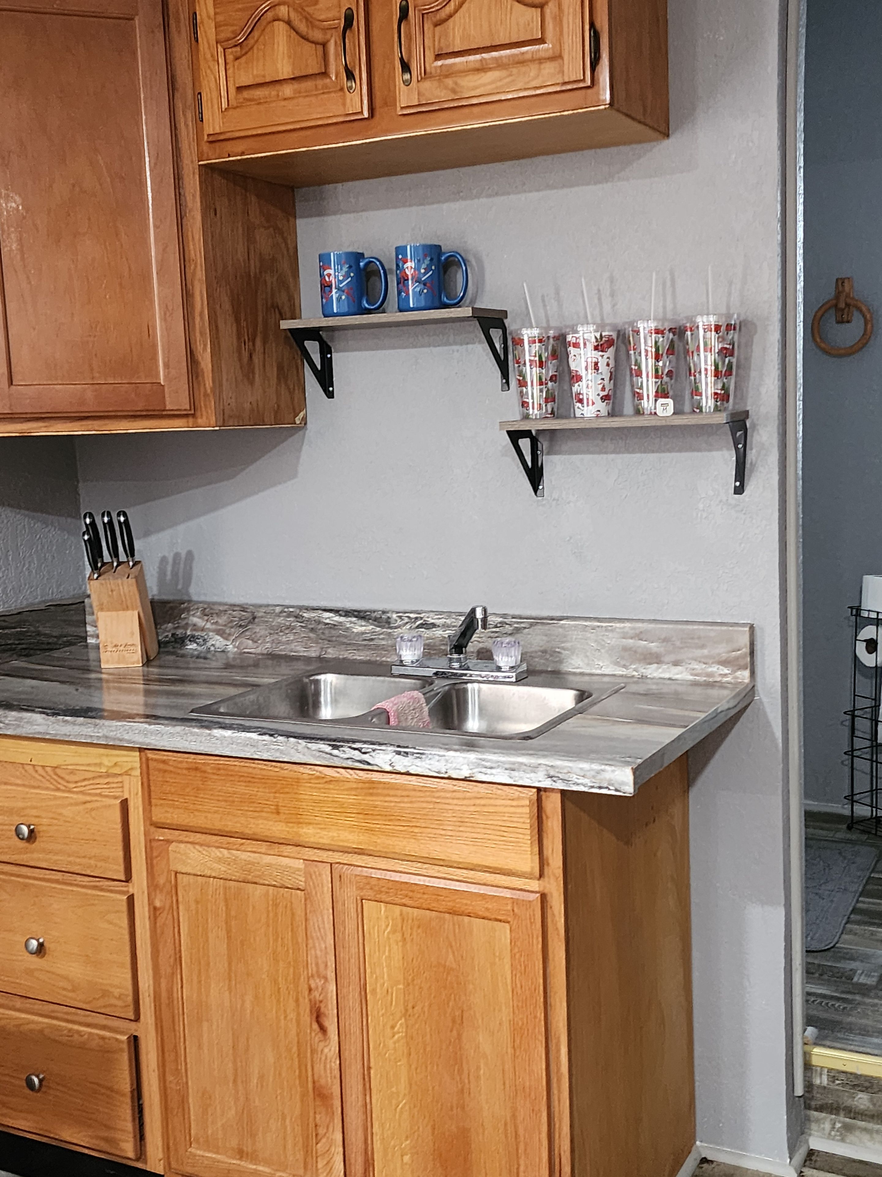 A kitchen with wooden cabinets, a marble countertop, a sink, and shelves with mugs and containers.
