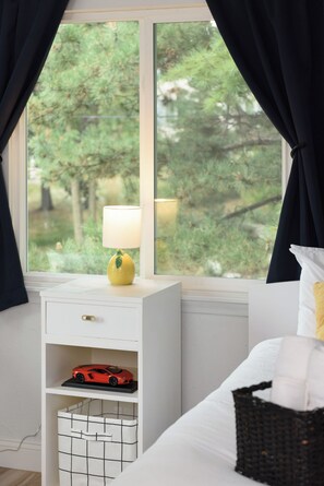 A bedroom with a bed, a nightstand with a lamp, a shelf with a toy car, and a window with curtains. - Lakefront 5BR Retreat on Lake Phalen-Sleeps 14-Fire Pit & Workspace (Saint Paul)