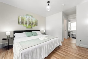 Une chambre moderne avec un grand lit, deux tables de chevet, un luminaire fixé au mur et une salle de bain visible.