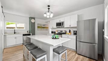 A modern kitchen with white cabinetry, a stainless steel refrigerator, a microwave, and a stove. There are four bar stools and a clock on the wall.