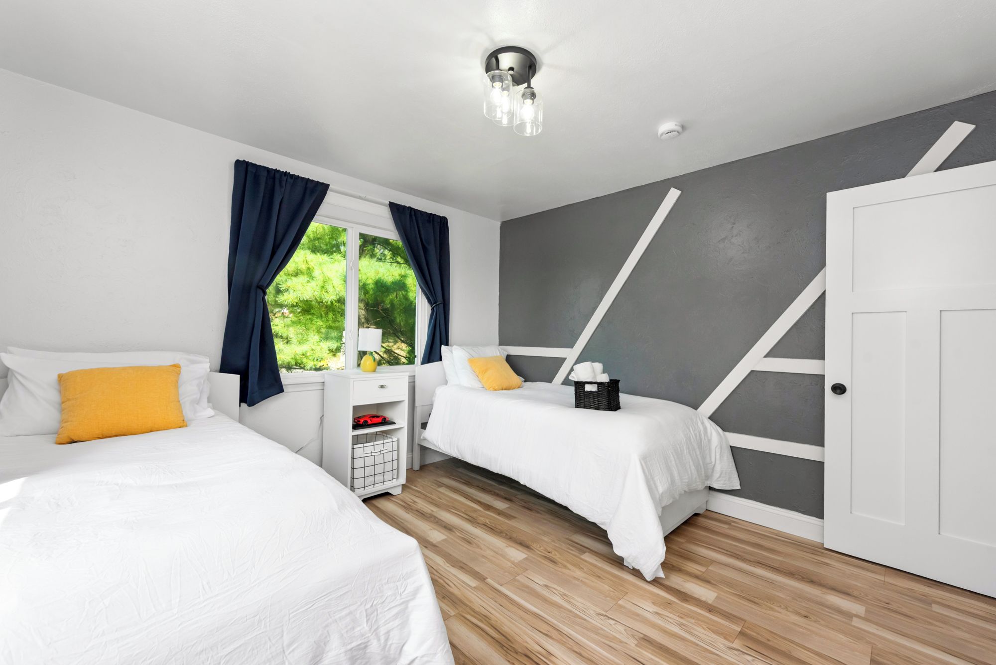 A bedroom with two beds, a window with curtains, a bedside table with a lamp, and a door.
