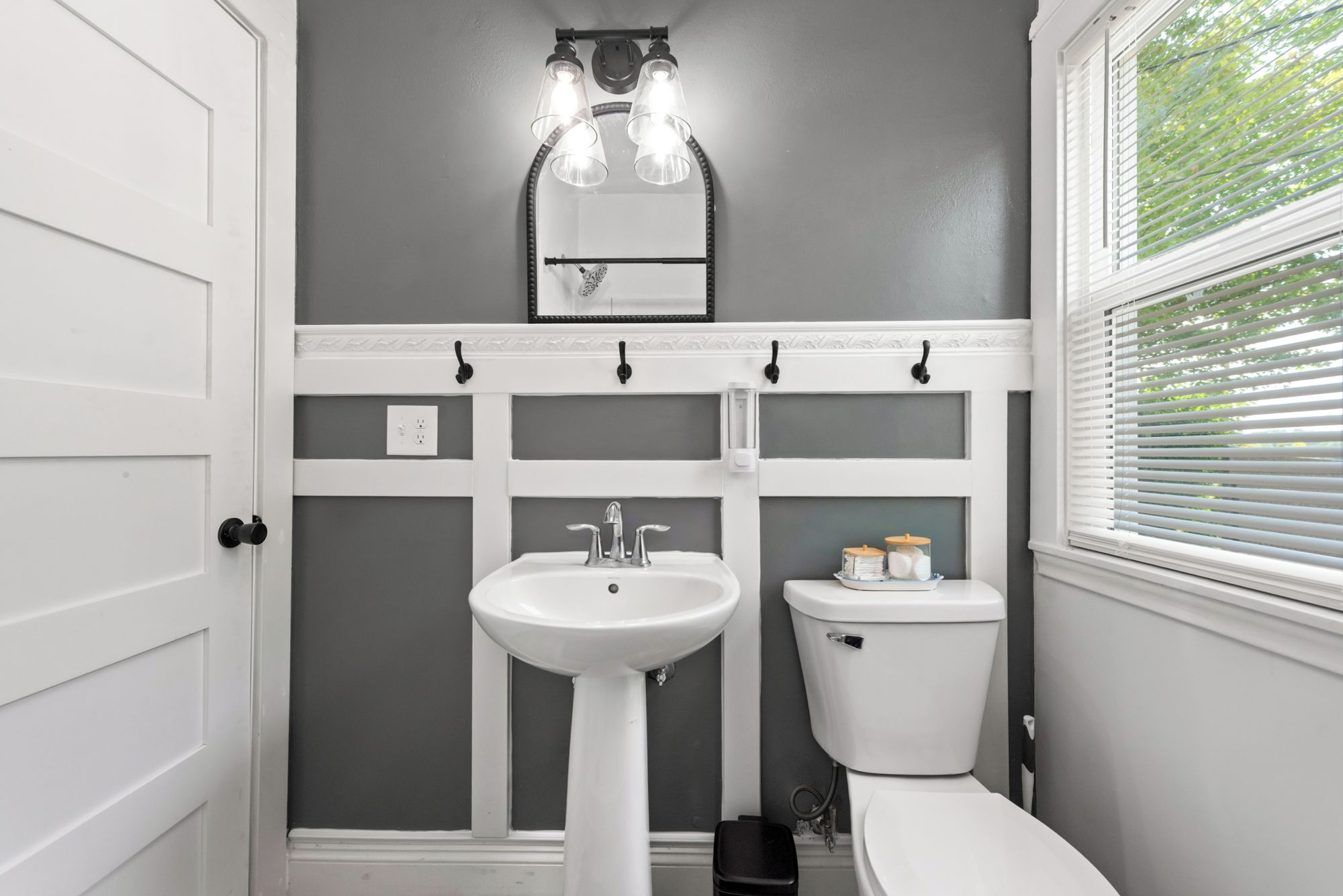 A bathroom with a white sink, toilet, and vanity. There is a mirror above the sink and a window with blinds on the right.
