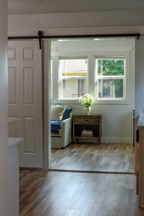 A room with a sliding door leading to an outdoor area, a window with a view of the outside, a chair with a patterned cushion, and a small wooden table with a vase of flowers.