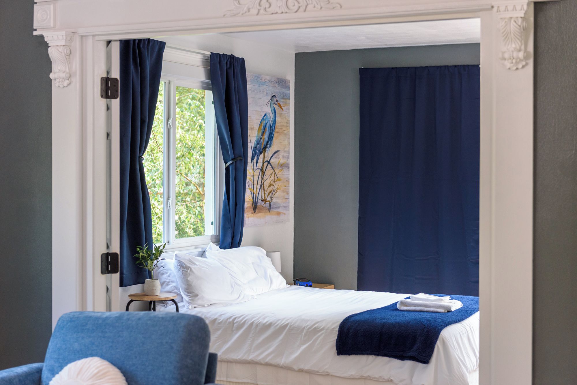 A hotel room with a bed, a chair, a small table with a plant, and a painting on the wall.