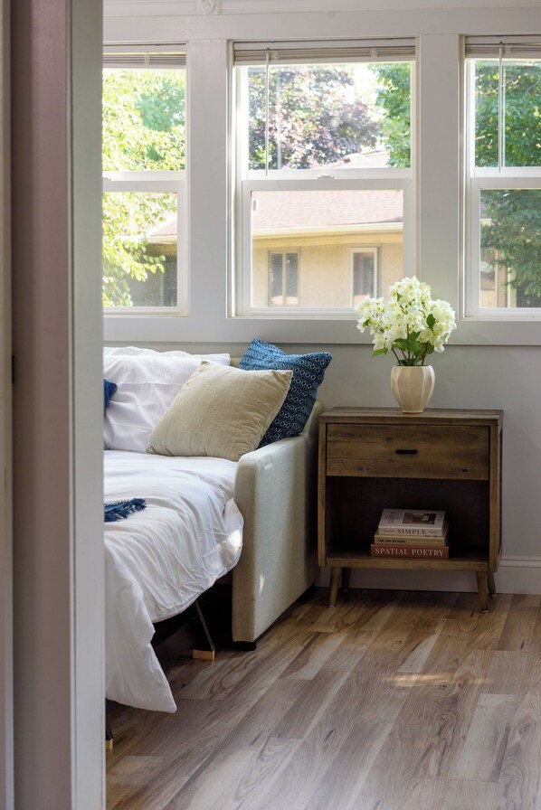 A bedroom with a bed, pillows, a bedside table with books, and a window with a view of greenery.