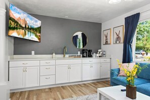A modern living room with a white cabinet, a coffee bar, a round mirror, and a blue sofa with a vase of flowers.