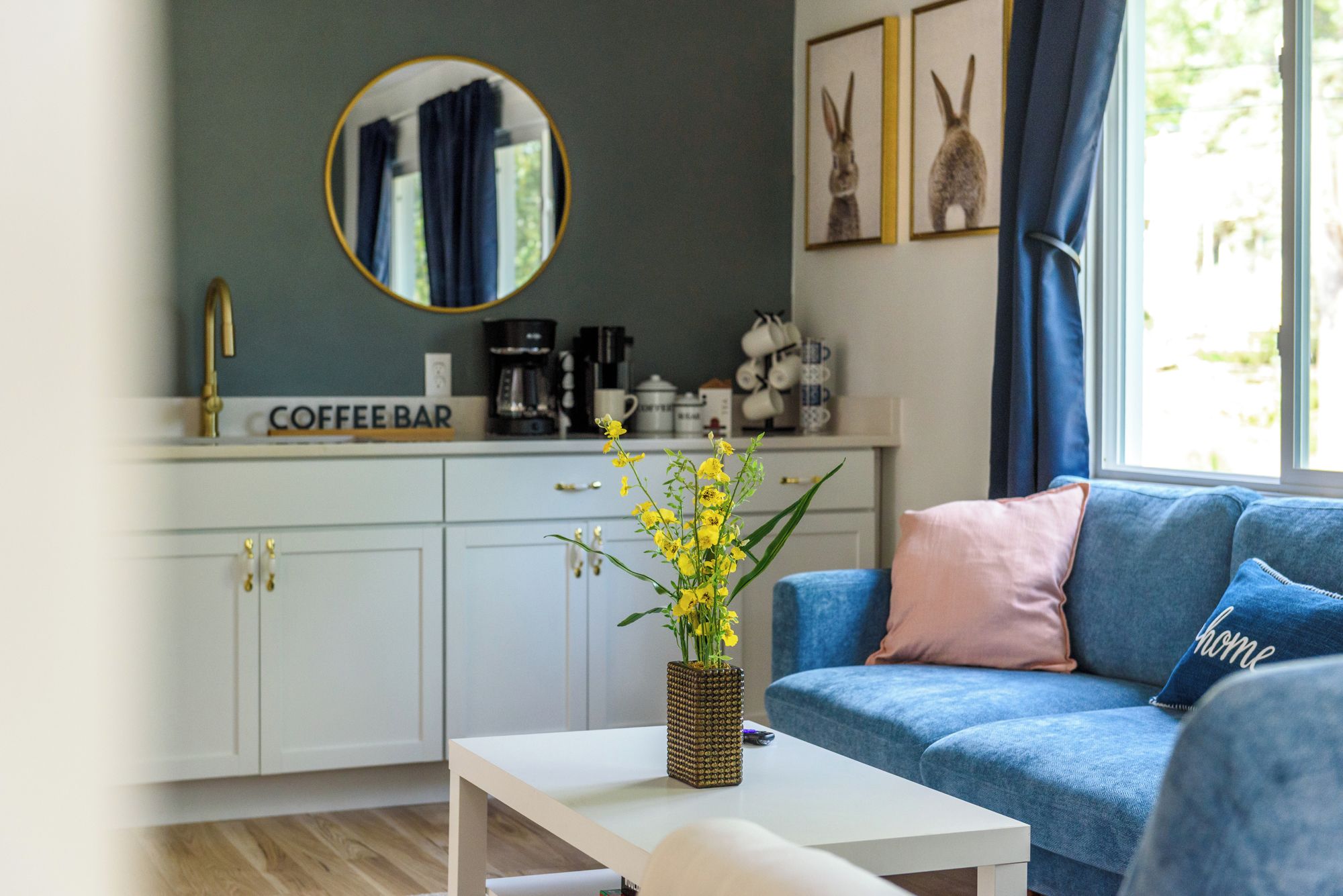 A modern living room with a blue sofa, a coffee bar in the background, and a round mirror above it.
