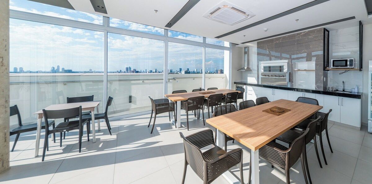 A modern office space with a large table and chairs, a kitchen area, and large windows offering a city view.