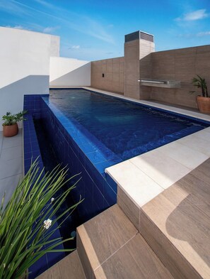 Piscina en la azotea con una fuente de agua azul cristalina, rodeada de baldosas blancas y beiges, y una planta en maceta a la derecha.