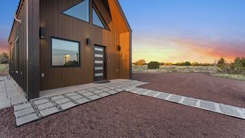 A modern two-story building with a sloped roof, large windows, and a gravel driveway leading to a wooden door.