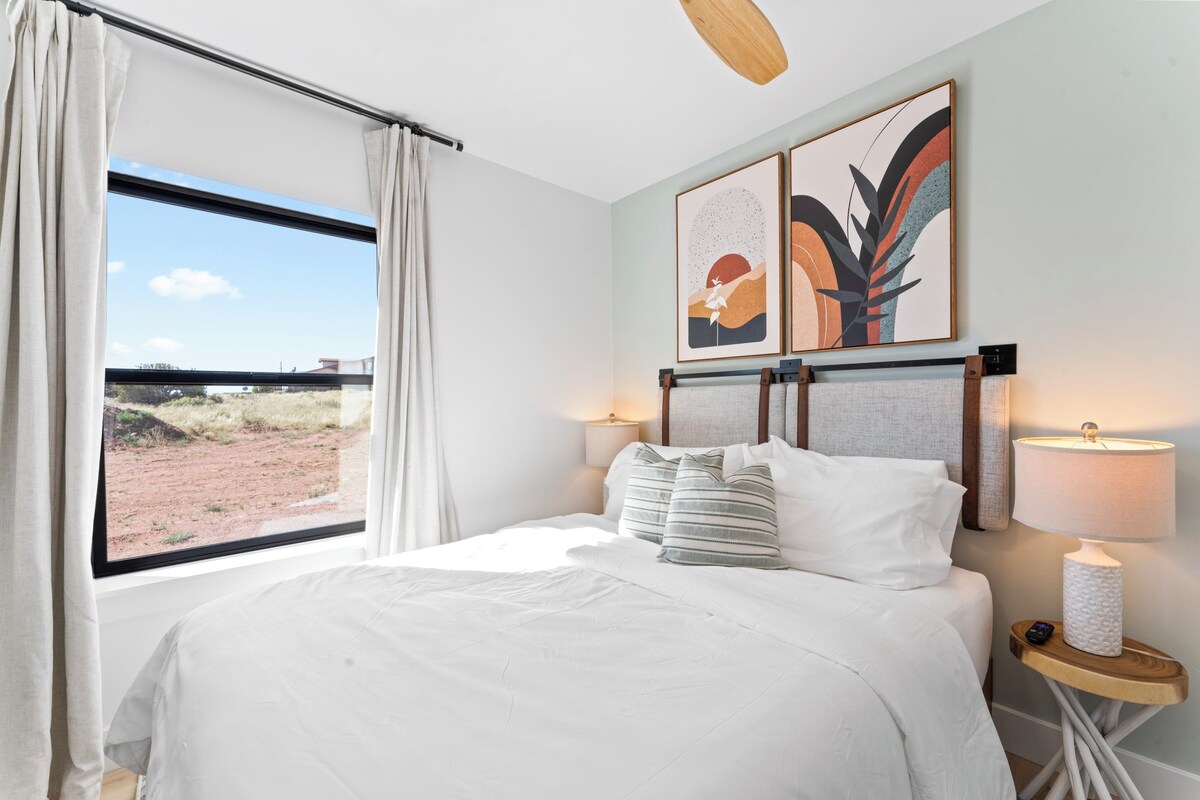Hotelkamer met een groot bed, een raam met gordijnen, twee kunstwerken aan de muur, een nachtkastje met een lamp en een plafondventilator.