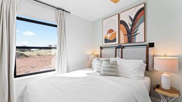 A hotel room with a large bed, a window with curtains, two wall-mounted artworks, a bedside table with a lamp, and a ceiling fan.