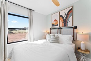 Hotelkamer met een groot bed, een raam met gordijnen, twee kunstwerken aan de muur, een nachtkastje met een lamp en een plafondventilator.