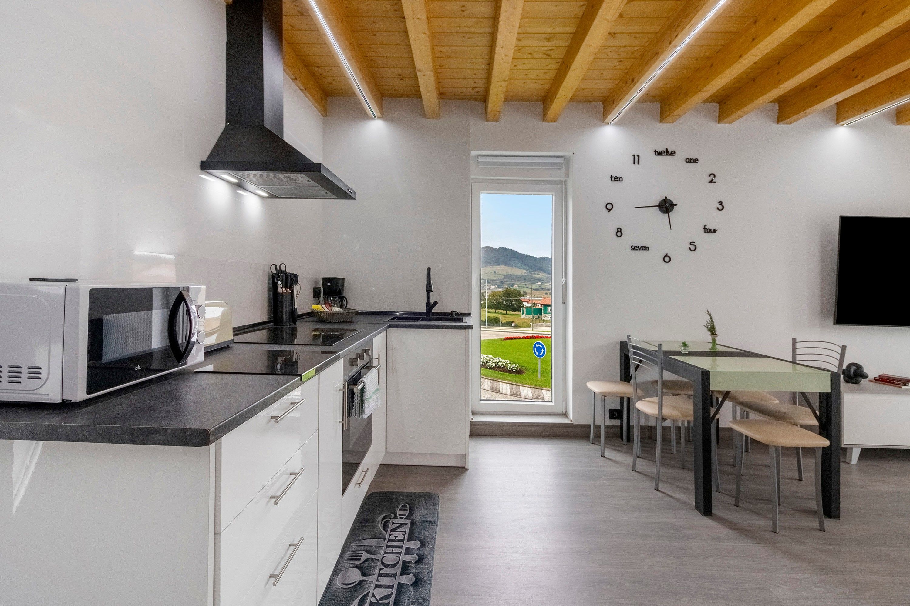 Een moderne keuken met een magnetron, broodrooster en koffiezetapparaat, een eethoek met tafel en stoelen, en een groot raam met uitzicht op een groen landschap.