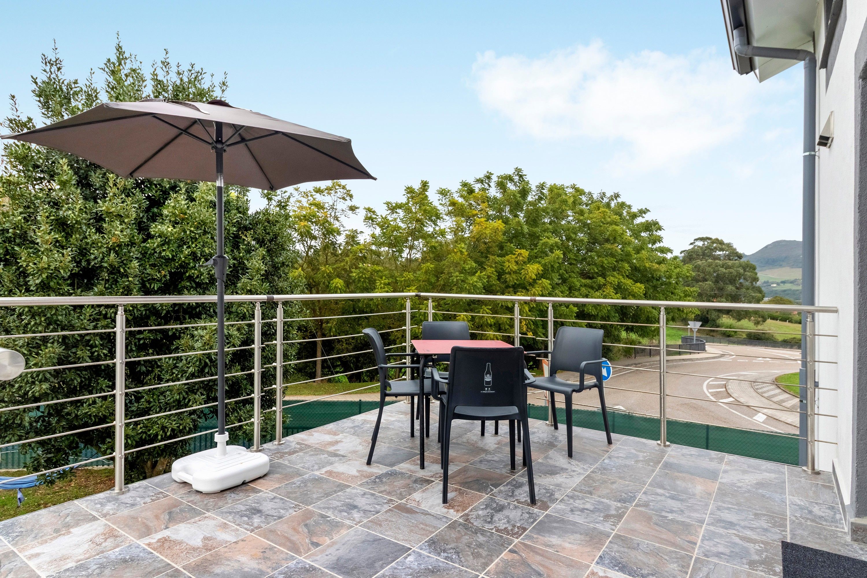Ein Balkon mit Tisch und Stühlen, einem großen Sonnenschirm und Blick auf eine Straße sowie Grünflächen.