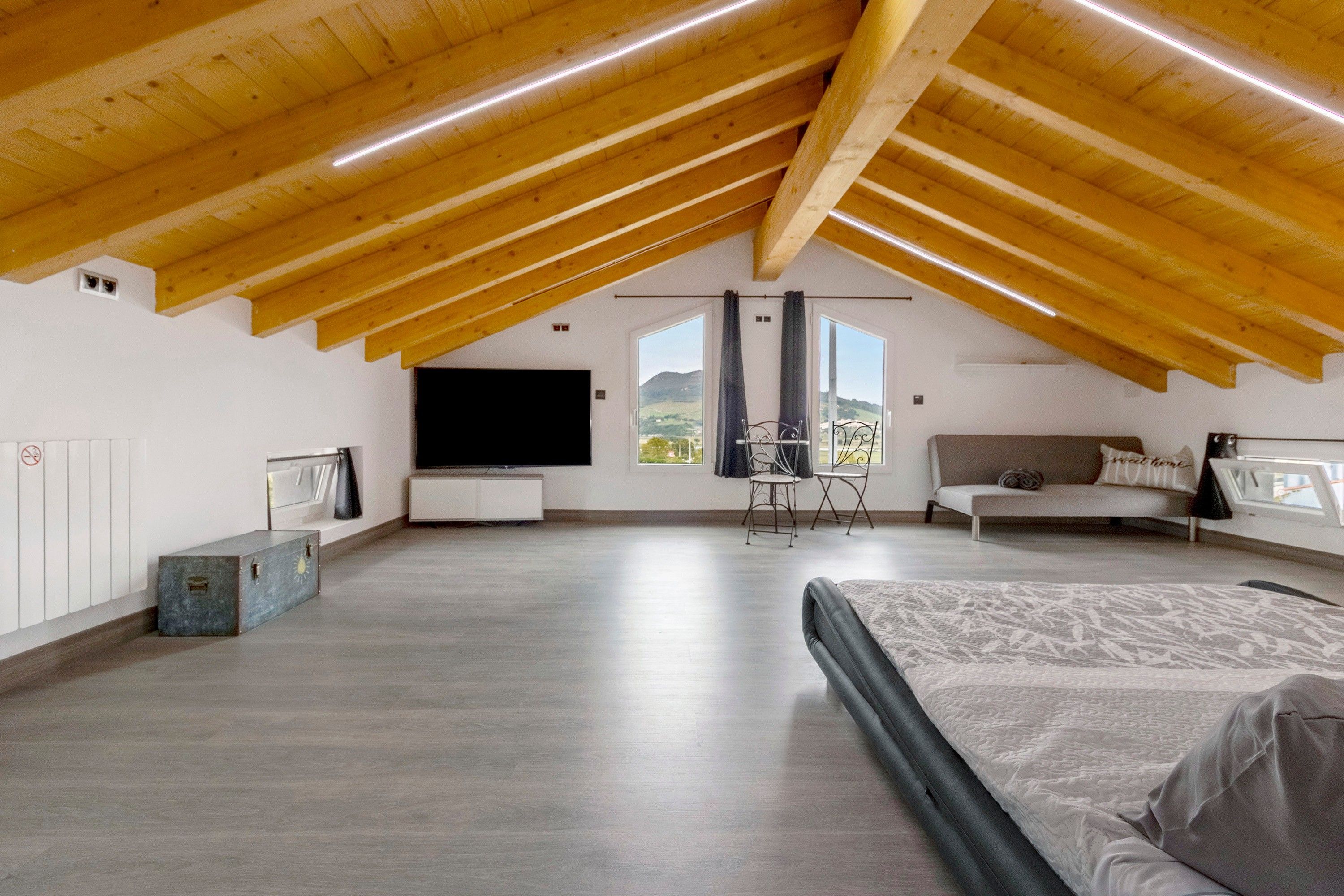 Amplia habitación en el ático con techo inclinado, cama, televisor, sofá y vistas al exterior.