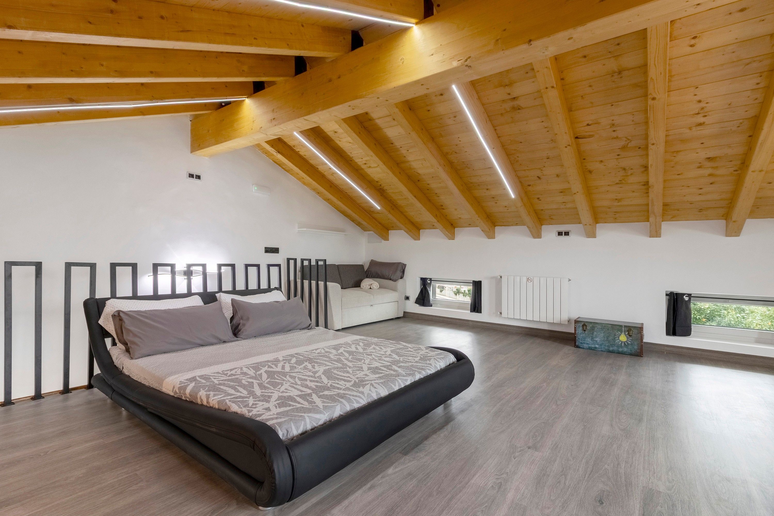 Ein modernes Schlafzimmer mit schrägem Dach, einem Bett mit gemustertem Überwurf und einem Fenster mit Aussicht. Ein Heizkörper ist ebenfalls vorhanden.