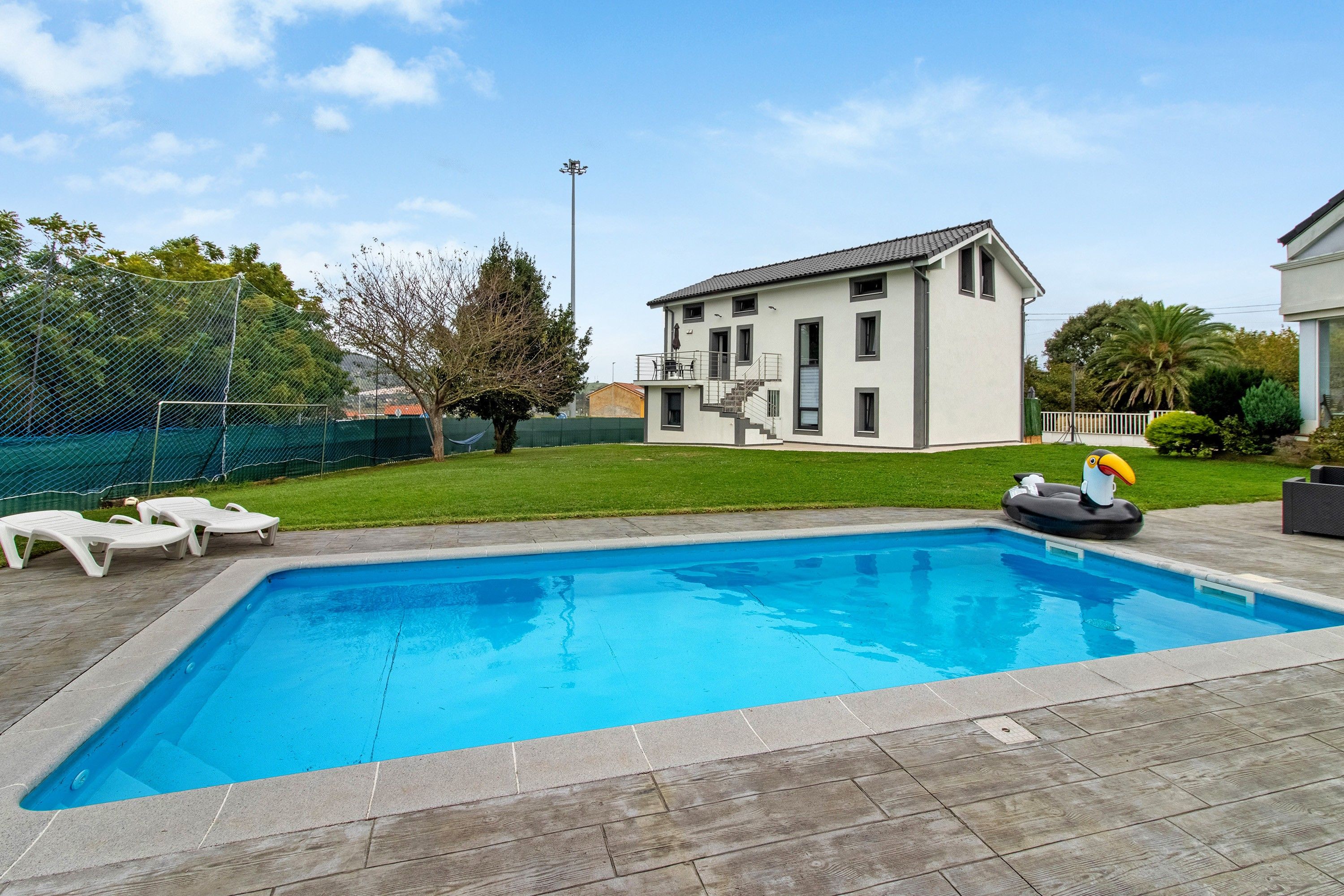 A rectangular swimming pool with a wooden deck surrounding it, a grassy lawn, and a two-story house with a balcony in the background.